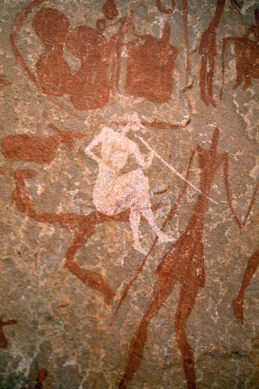 Tadrart, Algérie. Blanc homme assis avec les jambes croisées et portant une casquette, des vêtements et des chaussures pointues détient bâton (Instrument de musique?) à la bouche.