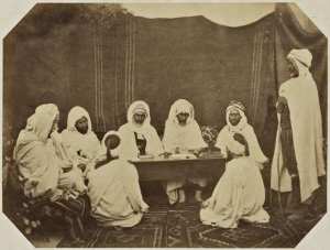 Midjelés de Mostaganem. Si Ahmed Bou Chenin, adel thaleb. Si Mohammed Ould El Bey Brahim bach-adel, thaleb alem. Si Ben Aouda, kadi de Mostaganem. Si Mohammed El Agachi, muphti 'président). Si Mohammed ben Kartaba, cadhi militaire. Si el Khatir, huissier, thaleb alem. Ahmed Bel Gassem sous-huissier, deux plaideurs (Image de propagande coloniale)