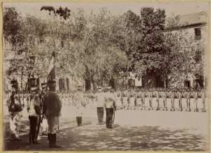 Remise de décorations. Quartier Blandan [Blida]. 14 juillet 1894 : capitaine Hoeberlé, capitaine Bajolle, lieutenant [porte drapeau] Féve, commandant Battréau, capitaine Rouchon, colonel Monthaulon, capitaine Hétet. (Image de propagande coloniale)