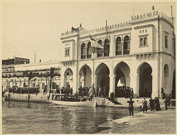 Arrivée de M. Loubet président de la République française. La vue est prise au moment où M. Loubet pose le pied sur la terre algérienne. Sous la voûte se trouvent les autorités algériennes, à gauche la musique des zouaves. L'amirauté ancienne construction, a eu la façade augmentée il y a quelques années. A gauche, on voit les bureaux de la Marine, la terrasse de l'Amirauté et le haut de la caserne construite sur la jetée Kheir-ed-dine. (Image de propagande coloniale)